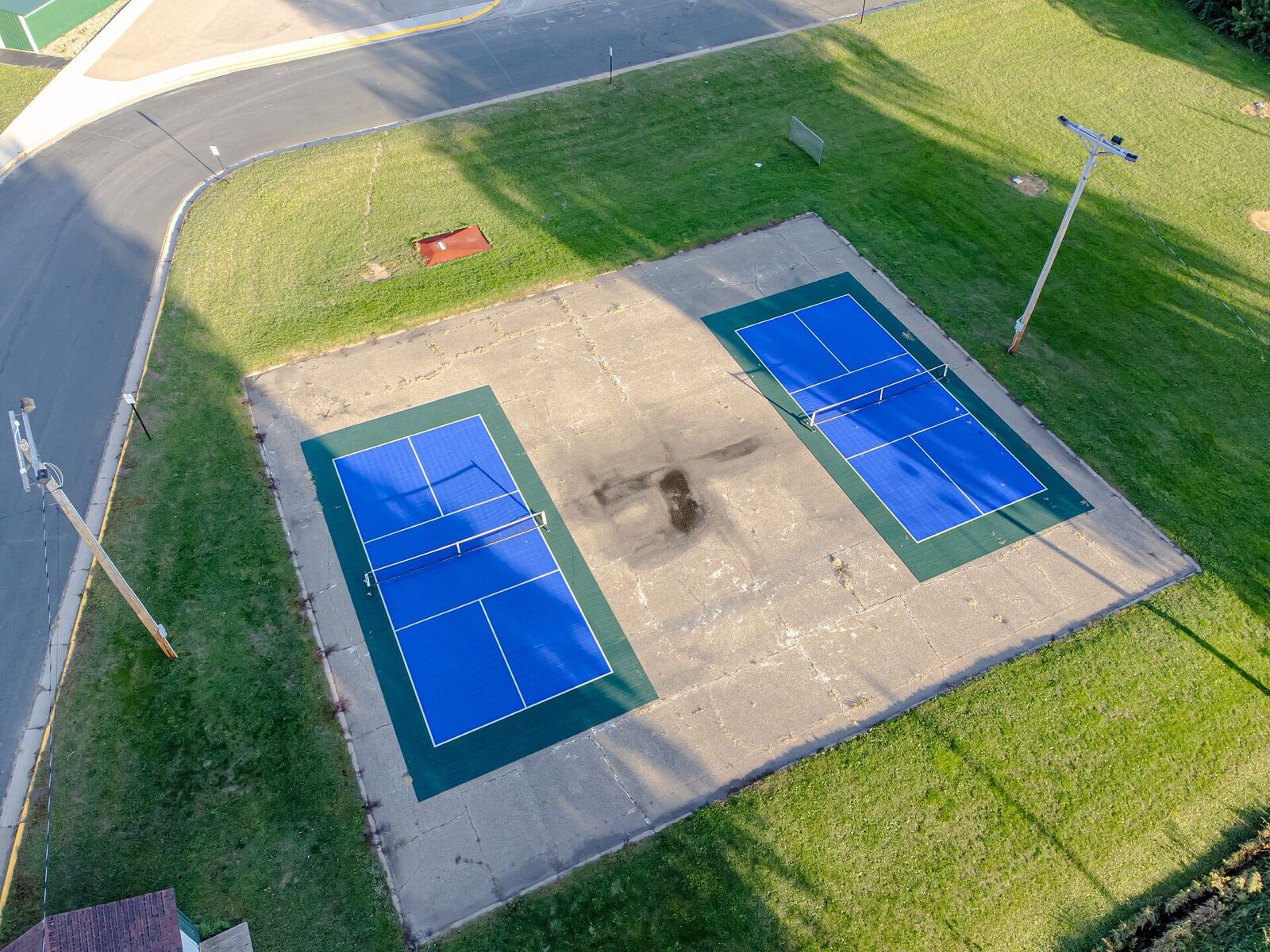 Two blue pickleball courts on a green surround, set in a concrete lot beside a lawn