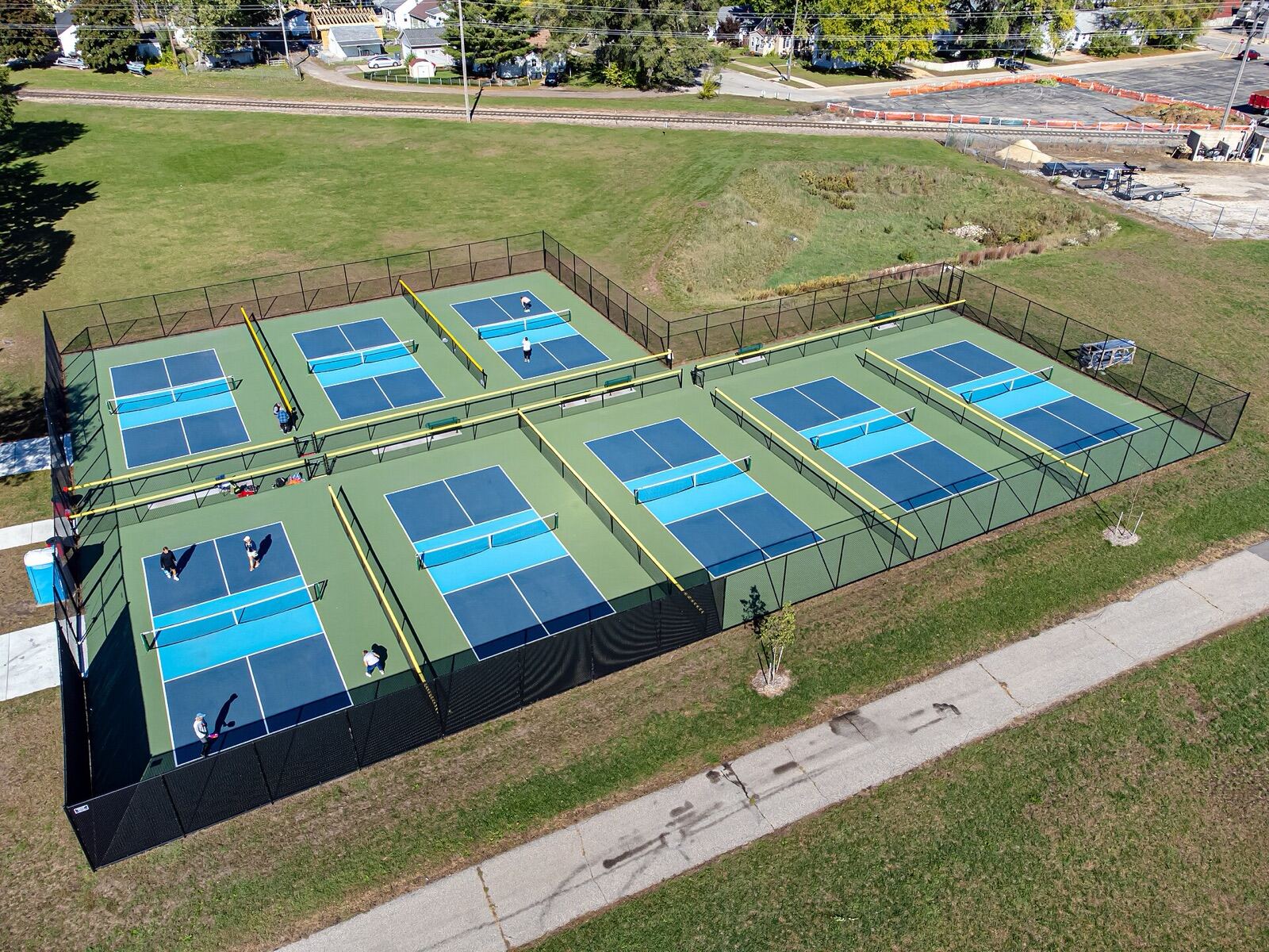 Aerial view of eight blue pickleball courts on a green surround inside a black fence, three in the back row and five in the front
